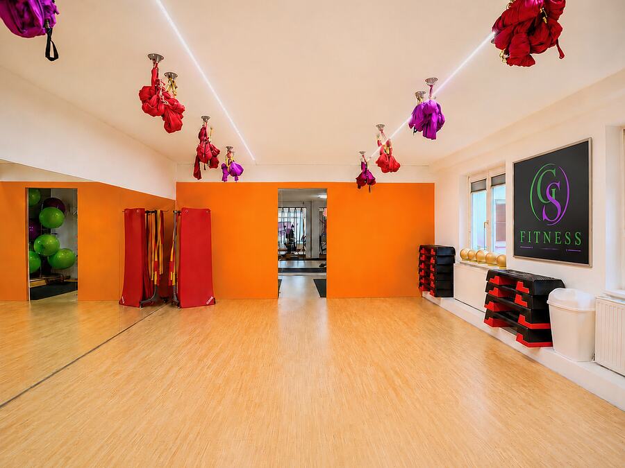 Gruppentrainingsraum von GS Fitness in Wien mit Holzboden, oranger Wand und Aerial-Yoga-Tuechern an der Decke